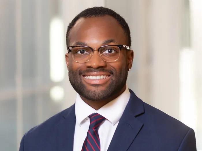 Uzochi Nwoko poses for a professional portrait wearing a navy suit, white shirt and striped tie against a softly lit indoor background.