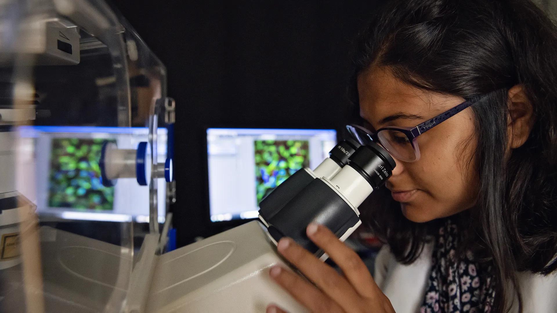 Female researcher with glasses looking into microscope with computer in background