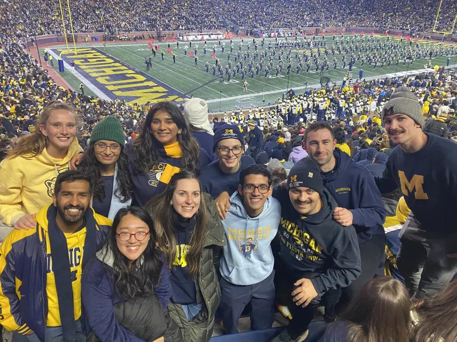 A group of approximately eleven students smiling together in the stands at a Michigan Stadium night game, wearing maize and blue University of Michigan apparel including hats, sweatshirts, and scarves. The marching band is performing on the field below, and the packed stadium crowd is visible in the background.